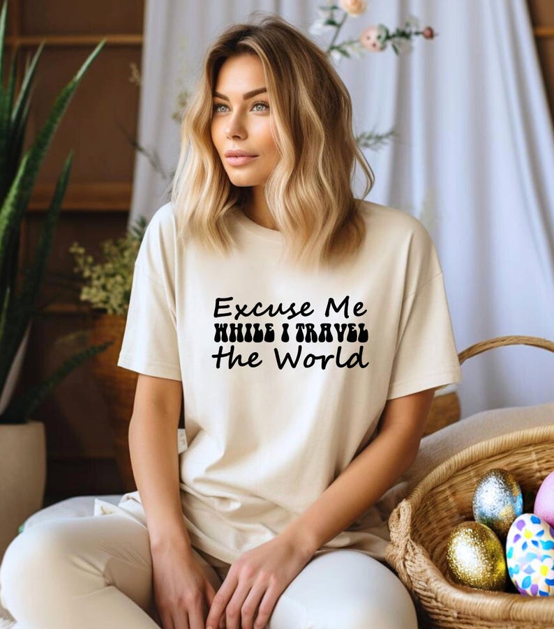 May include: A cream-coloured t-shirt with the text "Excuse Me While I Travel The World" in black. The shirt is paired with light-coloured trousers. A wicker basket with decorative Easter eggs is in the foreground.