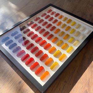 Puede incluir: Una exhibición enmarcada de objetos rectangulares coloridos dispuestos en filas. Los objetos cambian de color de azul a rojo, a naranja y a amarillo, con algunos objetos blancos al final. El marco es negro y el fondo es blanco.