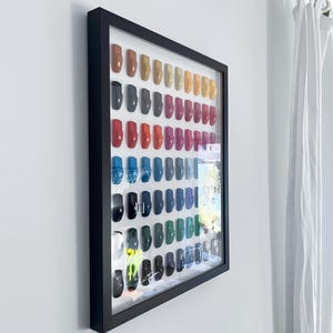 May include: A framed display of colorful nail polish bottles in various shades, including red, orange, pink, blue, and green. The bottles are arranged in rows within a black frame, creating a visually appealing collection. The frame is mounted on a white wall.