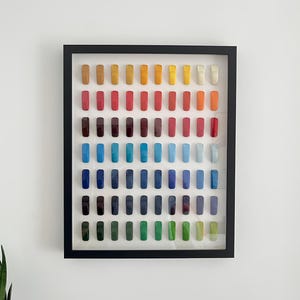 K&ouml;nnte beinhalten: Ein gerahmtes Kunstwerk mit einem Raster aus farbigen, rechteckigen Objekten. Die Anordnung zeigt ein Farbspektrum von warmen Gelb- und Oranget&ouml;nen bis hin zu k&uuml;hlen Blau- und Gr&uuml;nt&ouml;nen. Der Rahmen ist schwarz und das Kunstwerk h&auml;ngt an einer wei&szlig;en Wand.