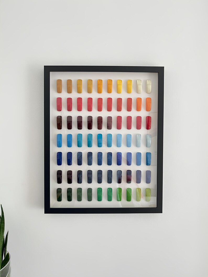 K&ouml;nnte beinhalten: Ein gerahmtes Kunstwerk mit einem Raster aus farbigen, rechteckigen Objekten. Die Anordnung zeigt ein Farbspektrum von warmen Gelb- und Oranget&ouml;nen bis hin zu k&uuml;hlen Blau- und Gr&uuml;nt&ouml;nen. Der Rahmen ist schwarz und das Kunstwerk h&auml;ngt an einer wei&szlig;en Wand.