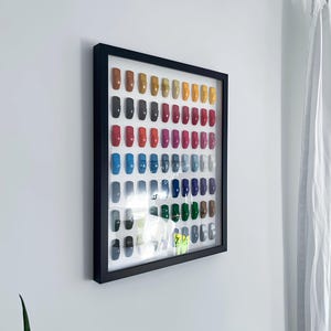 May include: A framed display of colorful artificial nails. The nails are arranged in rows and columns, showcasing a variety of shades including red, blue, green, and gold. The black frame is mounted on a white wall, with a curtain visible on the right.