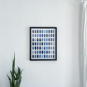 May include: A framed artwork featuring a grid of small blue squares in various shades, arranged in a gradient pattern. The artwork is displayed on a white wall.