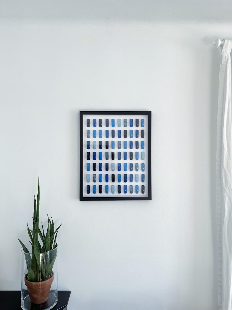 May include: A framed artwork featuring a grid of small blue squares in various shades, arranged in a gradient pattern. The artwork is displayed on a white wall.