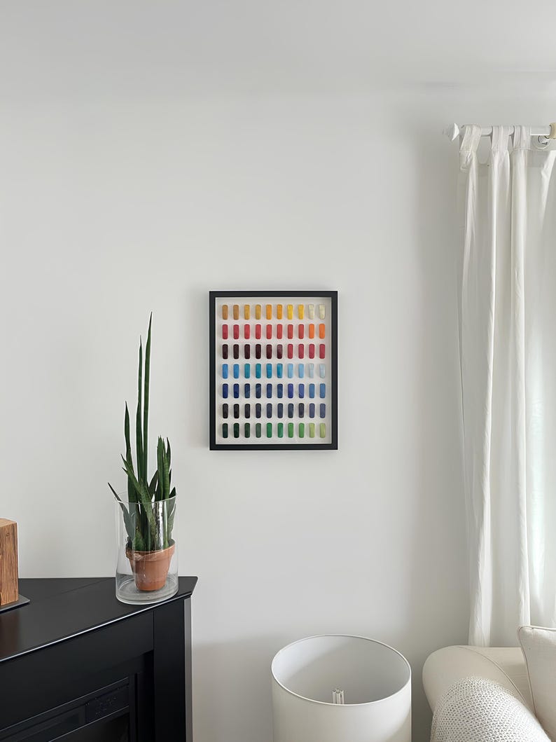 K&ouml;nnte beinhalten: Ein gerahmtes Kunstwerk mit einem Raster aus farbigen Rechtecken in einem Spektrum von Gelb bis Blau. Das Kunstwerk h&auml;ngt an einer wei&szlig;en Wand, mit einer Topfpflanze und einer Lampe im Raum.