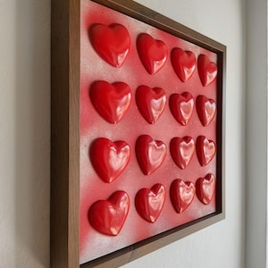 Könnte beinhalten: Ein gerahmtes Kunstwerk mit sechzehn glänzenden roten Herzformen, die auf einem rot lackierten Hintergrund angeordnet sind. Das Kunstwerk ist in einem dunkelbraunen Holzrahmen eingefasst. Das Stück ist an einer weißen Wand montiert.