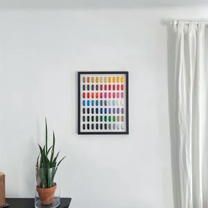 May include: A framed artwork featuring a grid of colorful spools of thread. The spools are arranged in rows and columns, showcasing a spectrum of colors from yellow to blue. The frame is black, and the artwork is displayed on a white wall.
