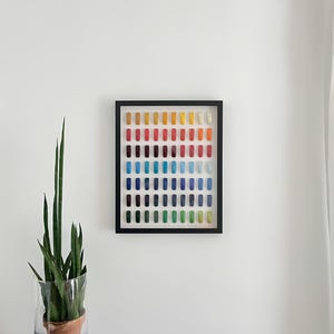 K&ouml;nnte beinhalten: Ein gerahmtes Kunstwerk mit einem Raster aus farbigen rechteckigen Objekten in Rot-, Orange-, Gelb-, Gr&uuml;n-, Blau- und Lilat&ouml;nen. Das Kunstwerk wird an einer wei&szlig;en Wand ausgestellt, mit einer Topfpflanze in einer Glasvase links.