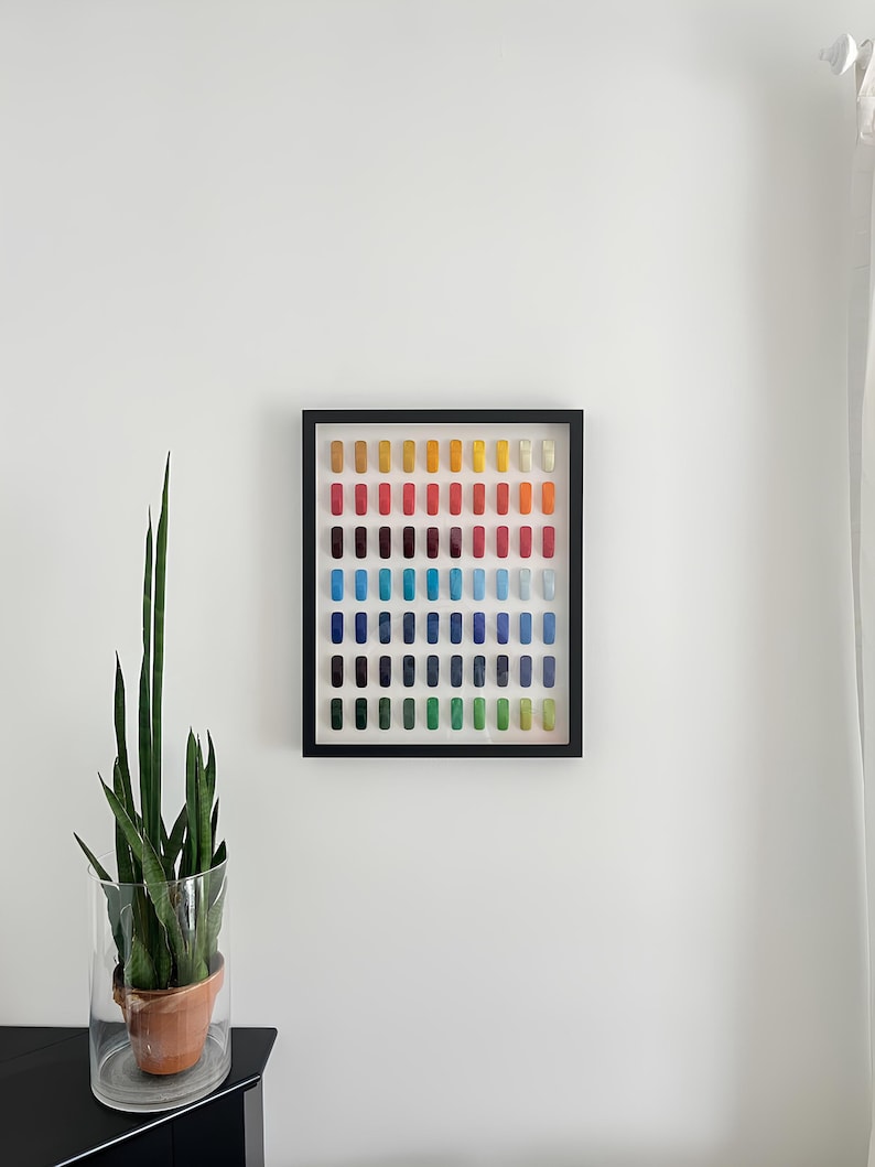 K&ouml;nnte beinhalten: Ein gerahmtes Kunstwerk mit einem Raster aus farbigen rechteckigen Objekten in Rot-, Orange-, Gelb-, Gr&uuml;n-, Blau- und Lilat&ouml;nen. Das Kunstwerk wird an einer wei&szlig;en Wand ausgestellt, mit einer Topfpflanze in einer Glasvase links.