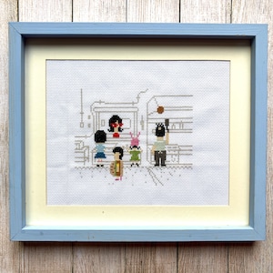 Op de afbeelding: Een ingelijst borduurwerk met kruissteek, met een familie van vijf cartoonfiguren. De vader draagt een zwart shirt en een zwarte hoed. De moeder draagt een rood shirt en een zwarte hoed. De kinderen dragen verschillende kleurrijke kleding. Het borduurwerk is op een witte achtergrond.