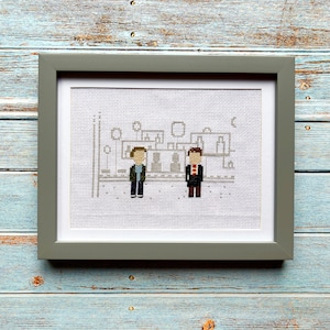 May include: A framed cross-stitch artwork depicting two pixelated figures standing in front of a building. The figures are wearing blue and red clothing. The background is a light gray color.