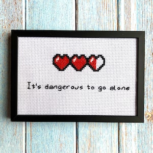 以下が含まれることがあります： ピクセル化されたハートの体力ゲージと「It's dangerous to go alone」というテキストが書かれた、額装されたクロスステッチ作品です。