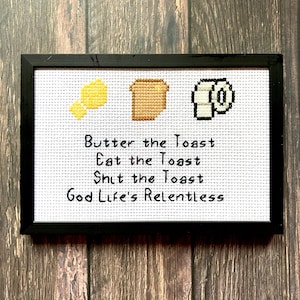 Peut inclure: Une broderie au point de croix blanche encadrée en noir avec un bâton de beurre jaune, une tranche de pain grillé brune et un rouleau de papier toilette. Le texte dit "Butter the Toast, Eat the Toast, Shit the Toast, God Life's Relentless."