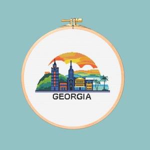 May include: A cross-stitch embroidery hoop featuring a pixelated cityscape with a sunset backdrop. The word "GEORGIA" is stitched in black at the bottom. The design includes buildings, palm trees, and a colorful sky.