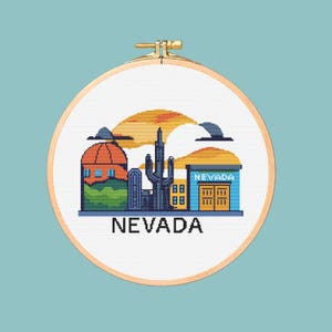 May include: A cross-stitch embroidery hoop featuring a pixelated landscape of Nevada. The design includes a building, cactus, and the word "NEVADA" in black. The background is a sunset with a teal-colored background.