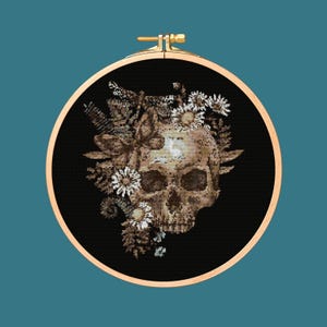 May include: A black and white cross-stitch embroidery hoop featuring a detailed skull with a floral wreath of white flowers and leaves.
