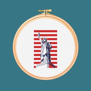 Peut inclure: Un cerceau de broderie en point de croix avec un fond blanc et une image rouge, blanche et bleue de la Statue de la Liberté. La Statue de la Liberté se tient sur une base avec un fond rayé rouge, blanc et bleu.