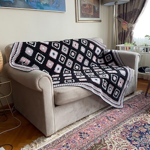 Könnte beinhalten: Eine schwarz-weiße Häkeldecke mit rosa Akzenten, die über einem beigen Sofa drapiert ist. Die Decke hat ein geometrisches Rautenmuster. Der Raum hat einen floralen Teppich, eine gewebte Lampe und kleine Topfpflanzen.