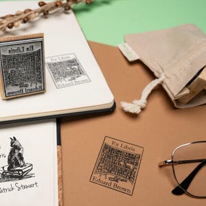 May include: A wooden stamp with the text "Ex Libris" and a library illustration, alongside a stamped image on a notebook. A canvas bag contains additional stamps. Also present are a pair of glasses and a sprig of berries.