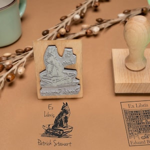 May include: Wooden stamps with illustrations and text, including a cat on a boat and a library scene. The stamps are on a brown surface with a light green background. A light green mug and decorative branches are also present.