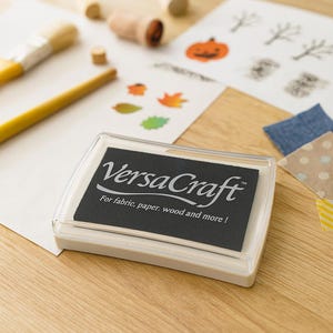 May include: A clear, rectangular VersaCraft ink pad with a black ink surface. The text "VersaCraft" and "For fabric, paper, wood and more!" are printed on the ink pad. The image also shows stamps, paintbrushes, and stamped leaf and pumpkin designs.