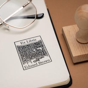 May include: A wooden stamp with a handle sits next to a notebook with the text "Ex Libris Eduard Brown" printed on it. Gold-rimmed glasses rest on the notebook. The stamp has a rectangular base and a rounded handle.