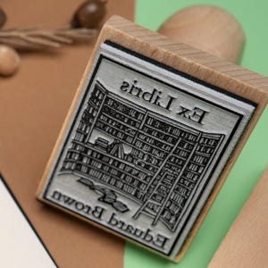 May include: A wooden stamp with a rectangular design featuring a library scene and the text "Ex Libris Edward Brown". The stamp is made of wood and has a detailed, engraved design. The background is a mix of brown and green.
