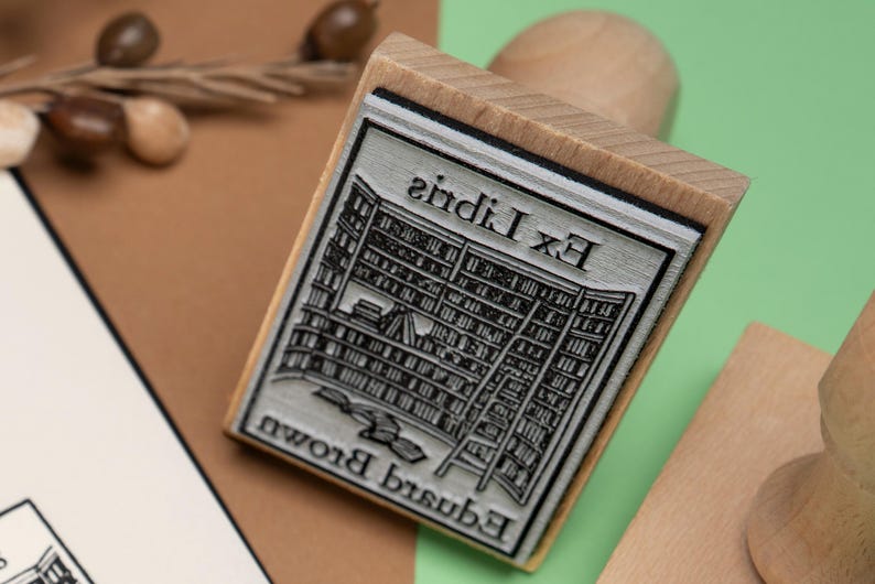 May include: A wooden stamp with a rectangular design featuring a library scene and the text "Ex Libris Edward Brown". The stamp is made of wood and has a detailed, engraved design. The background is a mix of brown and green.