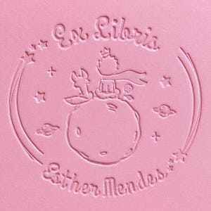 Op de afbeelding: Roze reliëf ontwerp met de tekst "Ex Libris" en "Esther Mendes". Het ontwerp bevat een klein figuur op een planeet, omringd door sterren en planeten. Het thema is literair en grillig.