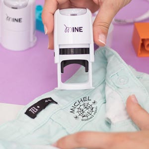 Puede incluir: Un sello blanco "mine" se utiliza para imprimir un diseño de telaraña negra, el nombre "MICHEL" y "GIMENEZ PERAL" en una camisa azul claro. El sello lo sostiene una persona. Otros elementos en el fondo incluyen un sello blanco y un bloque naranja.