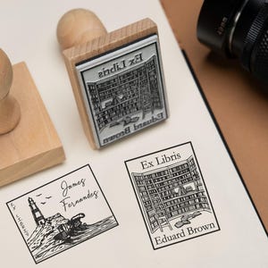 May include: Wooden stamps with black ink imprints of a lighthouse and a library. The stamps are on a white surface with a camera in the background. The text "Ex Libris" is visible on the stamps.