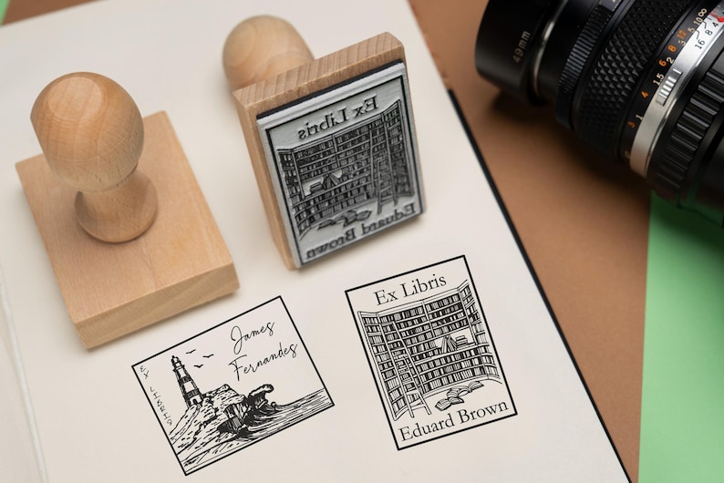 May include: Wooden stamps with black ink imprints of a lighthouse and a library. The stamps are on a white surface with a camera in the background. The text "Ex Libris" is visible on the stamps.