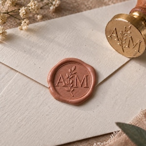 Op de afbeelding: Een rond, terracotta-kleurig waszegel met de letters A en M gescheiden door een takje met blaadjes, gedrukt op een crèmekleurige envelop. Daarnaast ligt een koperen waszegelstempel met een houten handvat, voorzien van hetzelfde A en M ontwerp.