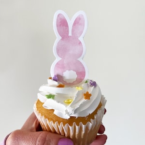 May include: A vanilla cupcake with white frosting and colourful sprinkles, topped with a pink watercolour bunny cutout. The cupcake is in a white paper case, ideal for Easter.