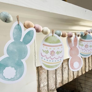May include: Easter garland with pastel-coloured egg and bunny cutouts. The garland includes decorative eggs and bunny shapes in shades of blue, pink, and yellow, ideal for Easter decorations. The garland is attached to a string with small wooden pegs.