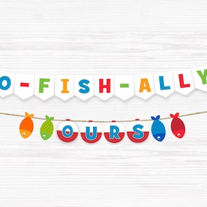 May include: A colourful banner reading "O-FISH-ALLY OURS". The banner features fish-shaped decorations in orange, green, blue, and red, strung on twine against a white wooden backdrop. The letters are in various colours.