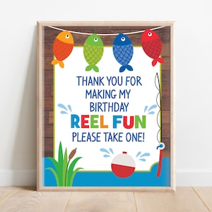 Może przedstawiać: Oprawiony wydruk o tematyce wędkarskiej. Wydruk przedstawia cztery kolorowe ryby wiszące na sznurku nad napisem "Thank you for making my birthday REEL FUN PLEASE TAKE ONE!" z grafiką związaną z wędkarstwem.