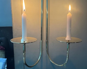 以下が含まれることがあります： 2本の腕を持つ金色の金属製の壁掛けキャンドルホルダー。各腕には丸いプレートと点灯した白いキャンドルが付いています。
