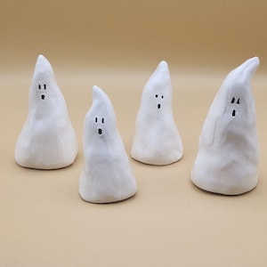 May include: Four small, white ceramic ghost figurines with simple black dot eyes and mouths. The ghosts have a rounded base and a pointed top, perfect for Halloween or spooky decor.