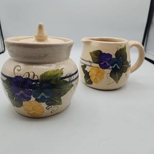 Könnte beinhalten: Ein Set aus zwei handbemalten Keramikbehältern für Zucker und Sahne. Die Behälter sind mit einem Blumendesign verziert, das lila und gelbe Blumen mit grünen Blättern zeigt. Die Behälter haben einen cremefarbenen Hintergrund mit einem blauen Streifen.