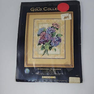 Puede incluir: Un kit de bordado crewel Dimensions The Gold Collection titulado Hydrangea Elegance. El empaque presenta un diseño floral de hortensias azules y moradas con hojas verdes sobre un fondo beige, que mide 28 cm por 36 cm.