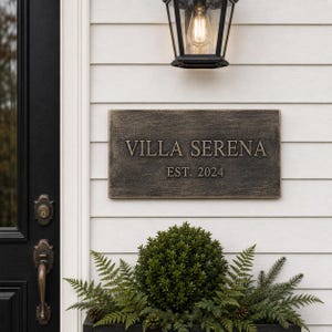 May include: A rectangular, dark bronze-toned house plaque with a weathered, textured finish. The plaque features the embossed text VILLA SERENA in a serif font, with the text EST. 2024 positioned directly below in a smaller, matching font style.