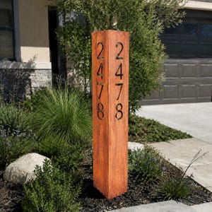May include: A tall, square, rust-colored metal address post featuring vertical cutouts of the numbers 2, 4, 7, and 8 on two visible sides. The pillar has a weathered, oxidized finish and stands upright as a functional outdoor house number display.