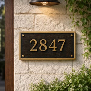 May include: A rectangular house number plaque featuring the digits 2847 in a metallic gold finish. The plaque has a dark, textured background with a gold-toned frame and is secured at each corner with small, circular gold-colored metal fasteners.