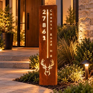 May include: A tall, rectangular, rust-colored metal address sign with vertical backlighting. The sign displays the numbers 27861, the text County Road H8, and a cutout graphic of a deer head at the bottom, all illuminated from within.