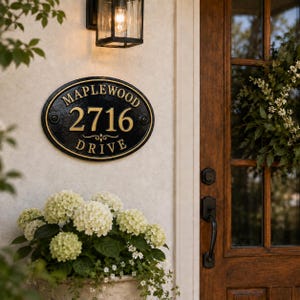 May include: An oval black house address plaque with gold lettering reading MAPLEWOOD 2716 DRIVE. The plaque features a raised border and two mounting screws on the sides, mounted on a light-colored wall next to a wooden door and a light fixture.