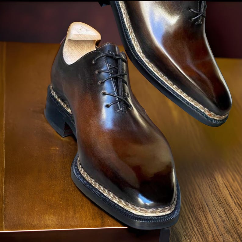 Puede incluir: Zapatos de vestir de cuero marr&oacute;n con acabado brillante. Los zapatos tienen cordones negros, suela negra gruesa y borde cosido decorativo. Un horma de madera est&aacute; dentro de un zapato.