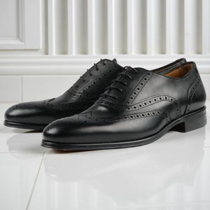May include: A pair of black leather dress shoes with brogue detailing. The shoes have a classic wingtip design with decorative perforations and a lace-up closure. The shoes are displayed on a white surface.