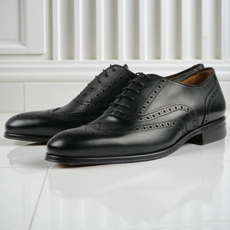 May include: A pair of black leather dress shoes with brogue detailing. The shoes have a classic wingtip design with decorative perforations and a lace-up closure. The shoes are displayed on a white surface.