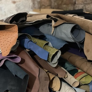 K&ouml;nnte beinhalten: Ein Haufen verschiedener Lederh&auml;ute in Braun-, Blau-, Gr&uuml;n- und Lilat&ouml;nen. Einige H&auml;ute sind gerollt, andere gefaltet, wodurch verschiedene Texturen und Muster entstehen. Das Leder scheint von hoher Qualit&auml;t zu sein.